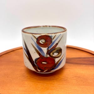 May include: A small ceramic cup with a cream-colored base featuring hand-painted floral motifs in deep red, brown, and blue. The rim is finished with a thin brown band, and the exterior surface displays a speckled texture throughout the clay body.