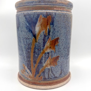 May include: A cylindrical ceramic utensil holder featuring a mottled blue and grey glaze with a decorative brown, plant-like pattern on the front. The vessel has a raised rim at the top and a distinct base, both finished in a warm, earthy brown tone.