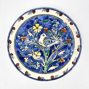 May include: A round ceramic plate featuring a hand-painted design of a white bird perched on a green branch surrounded by yellow, blue, and orange flowers against a deep blue background, with a cream-colored border with brown accents.
