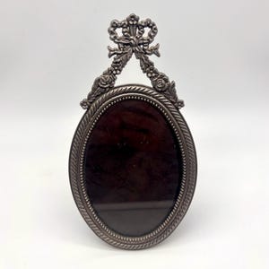 May include: An oval-shaped metal picture frame with a dark, reflective center. The frame features a rope-textured border and an ornate top ornament depicting a bow and floral details. The metal has a muted, antique silver-toned finish throughout.