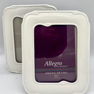 May include: Two white ceramic photo frames with a rectangular shape and curved edges. The frames have a 5x7 inch opening and the text "Allegro" and "Photo Frame 5x7" are visible.