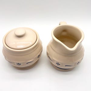 May include: A cream-colored ceramic sugar bowl with a matching lid and a cream-colored ceramic creamer pitcher. Both pieces feature a dark blue floral pattern around the base and a smooth, glossy finish on the exterior and interior surfaces.