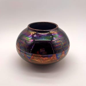 May include: A round, squat ceramic vase with a wide opening and a smooth, reflective surface. The finish features an iridescent, metallic glaze that shifts between deep purple, green, and copper tones, with a distinct horizontal band of orange.