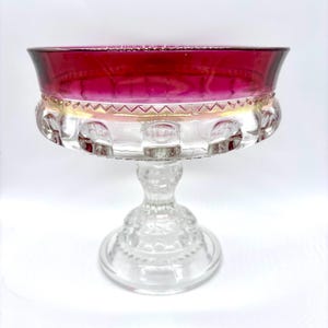 May include: A clear glass pedestal bowl with a vibrant red rim. The bowl features a decorative pattern around the base and a detailed stem. The rim transitions from red to a golden hue. This elegant piece is suitable for serving desserts or displaying decorative items.
