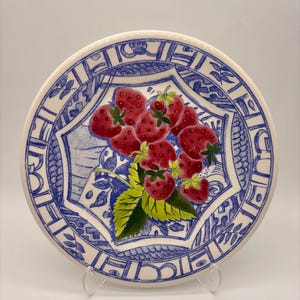 May include: A round ceramic plate featuring a central cluster of red strawberries with green leaves. The plate is decorated with a blue and white border pattern that includes intricate line work and stylized geometric shapes surrounding the fruit.