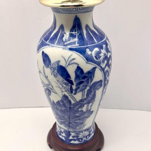 May include: A blue and white porcelain vase lamp featuring intricate hand-painted botanical and bird motifs. The lamp has a polished gold-tone metal lid at the top and rests on a dark brown circular wooden base, showcasing a classic ceramic design.