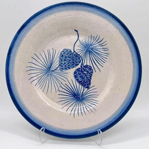 May include: A round ceramic plate with a blue rim and a cream-colored center. The plate features a blue illustration of pine cones and stylized pine needles. The plate is supported by a clear plastic stand.