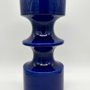 May include: A cobalt blue ceramic vase with a unique hourglass shape. The vase has a wide base and top, connected by a narrower, cylindrical midsection. The glossy finish reflects light, highlighting its smooth curves.
