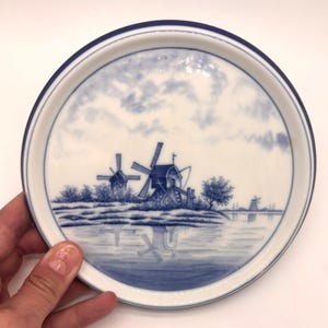 May include: A round ceramic plate featuring a blue and white transferware design of a Dutch landscape with windmills, trees, and a body of water. The plate has a solid dark blue rim and a white background with soft blue clouds in the sky.