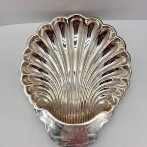 May include: A polished silver-toned metal dish shaped like a large scallop shell. The dish features deep, radiating fluted grooves that extend from the base to the scalloped edge, creating a reflective surface with a smooth, flat handle area.