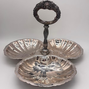 May include: A silver-plated three-section serving dish featuring three scalloped, shell-shaped bowls arranged around a central vertical post. The post is topped with an ornate, circular handle decorated with intricate scrollwork and leaf motifs.