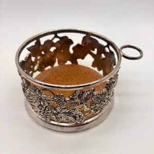 May include: A circular silver-toned metal coaster holder featuring an intricate openwork design of grape clusters and vine leaves. The base of the holder is lined with a light brown cork insert, and a small circular handle extends from one side.