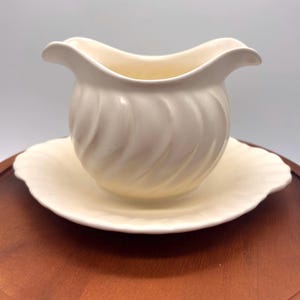 May include: Cream-colored gravy boat with a scalloped rim, resting on a matching saucer. The boat has a curved handle and a spiral design on the exterior. The set is made of ceramic.