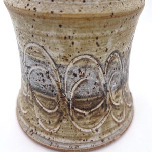 May include: A cylindrical ceramic vessel with a speckled tan and brown glaze. The body features a horizontal band of carved, overlapping circular patterns that reveal a lighter gray tone beneath the darker, textured surface of the clay.