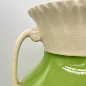 May include: A ceramic pitcher with a cream-colored top and handle, and a bright green body. The top has a textured, ribbed design, and the handle curves gracefully from the rim. The pitcher is likely vintage or antique.