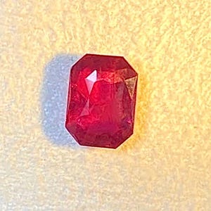 May include: A faceted, rectangular-cut ruby gemstone. The gem is a deep red color with multiple facets that reflect light. The gemstone is set against a textured, off-white background.
