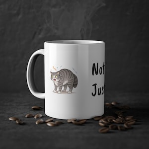 Peut inclure: Mug en céramique blanche avec un chat de dessin animé surpris et le texte "Not Just". Le chat est gris et blanc avec des yeux jaunes. La tasse a une anse noire et est entourée de grains de café.