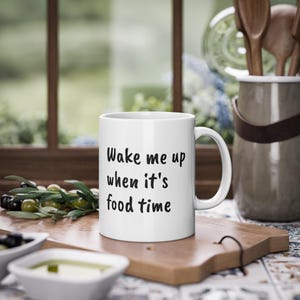 Könnte beinhalten: Weiße Keramiktasse mit dem Text "Wake me up when it's food time" in Schwarz. Die Tasse steht auf einem Holzschneidebrett mit Oliven und einem Behälter mit Olivenöl im Vordergrund. Im Hintergrund ist ein Fenster.