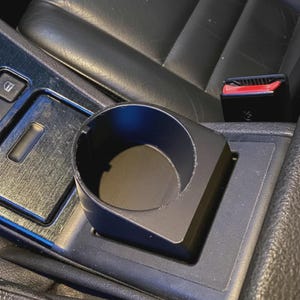 Op de afbeelding: Een matzwarte kunststof bekerhouder-inzetstuk met een cilindrische opening en een rechthoekige basis, ontworpen voor de middenconsole van een voertuig. Het accessoire heeft een gladde afwerking en een inkeping in de rand voor bekers.