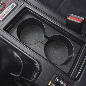 May include: A black automotive center console featuring two circular cup holders, a gear shift boot, and a control panel with buttons labeled C.DIFF, AUTO, and seat heater icons with HI and LO settings, alongside a red seatbelt release button.