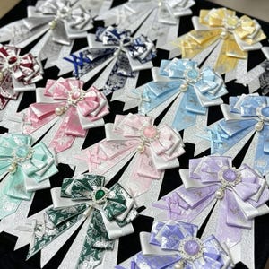 May include: A collection of multi-colored ribbon bow brooches featuring layered fabric, intricate floral patterns, and central decorative cabochon stones surrounded by small rhinestones, each accented with a single dangling faux pearl bead.