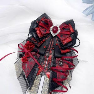 May include: A gothic-style hair accessory featuring a large black bow with red patterned fabric and a central red jewel. The design includes black chains, red ribbons, and small cross charms, creating a dramatic and ornate look.