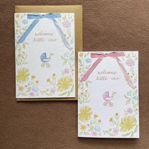 Pode incluir: Dois cartões de felicitações brancos com o texto "welcome little one" em cursivo. Um cartão apresenta uma fita azul e uma ilustração de carrinho de bebé azul, enquanto o outro tem uma fita rosa e um carrinho rosa. Ambos têm motivos florais.