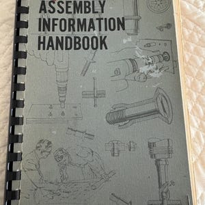 Puede incluir: Un manual gris de "Assembly Information Handbook" con encuadernación en espiral negra. La portada presenta el título en letras negras grandes, ilustraciones de herramientas y una firma roja. El manual parece ser vintage.
