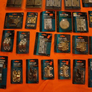 May include: An assortment of hardware items packaged in plastic blister packs. The items include hinges, door stops, sash locks, tension springs, and chair leg braces. The packages are labelled with the product name and brand.
