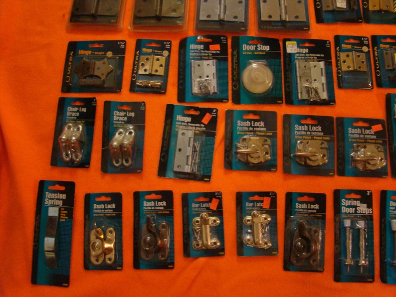 May include: An assortment of hardware items packaged in plastic blister packs. The items include hinges, door stops, sash locks, tension springs, and chair leg braces. The packages are labelled with the product name and brand.