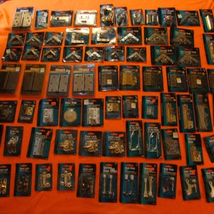 May include: A selection of door hardware in individual packages displayed on a bright orange surface. The hardware includes hinges, door stops, door latches, and corner braces. The packages are labelled with the type of hardware and the brand name.