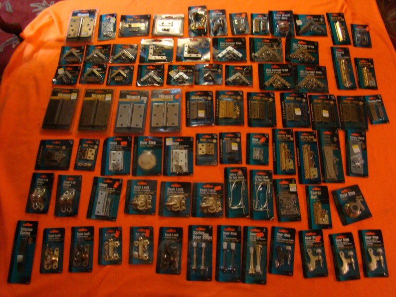 May include: A selection of door hardware in individual packages displayed on a bright orange surface. The hardware includes hinges, door stops, door latches, and corner braces. The packages are labelled with the type of hardware and the brand name.