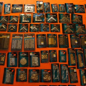 May include: A selection of hardware items, including hinges, door stops, barrel bolts, and sash locks, are displayed on a bright orange surface. The items are packaged in clear plastic bags with white labels and black text. The labels include the words "Hinges", "Door Stop", "Barrel Bolt", "Sash Lock", and "ULTRA".