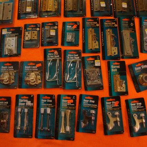 May include: A selection of door hardware in a variety of finishes, including door stops, sash locks, barrel bolts, hinges, and wire coat hooks. The hardware is packaged in clear plastic bags with labels and prices.