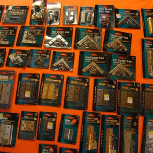 May include: A selection of hardware items, including hinges, door stops, corner braces, and barrel bolts, are displayed on a bright orange surface. The items are packaged in clear plastic bags with labels that include the product name and brand. The items are arranged in rows and columns, creating a visually appealing display.