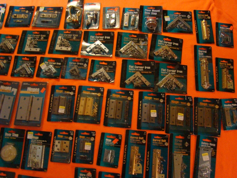 May include: A selection of hardware items, including hinges, door stops, corner braces, and barrel bolts, are displayed on a bright orange surface. The items are packaged in clear plastic bags with labels that include the product name and brand. The items are arranged in rows and columns, creating a visually appealing display.