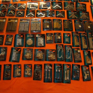 May include: An assortment of door hardware in individual packaging. The hardware includes hinges, door stops, door knobs, and other hardware. The packaging is clear plastic with black and blue lettering. The hardware is made of metal and is in various finishes, including brass, nickel, and chrome.