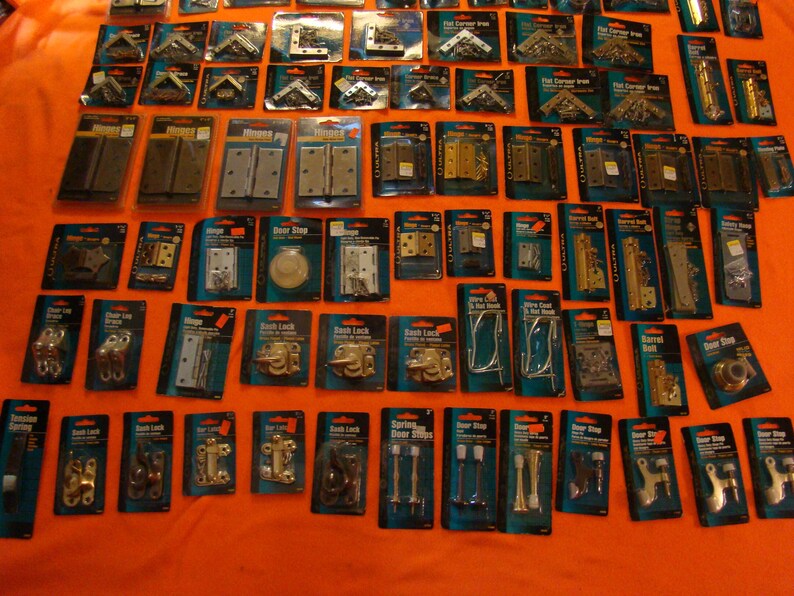May include: An assortment of door hardware in individual packaging. The hardware includes hinges, door stops, door knobs, and other hardware. The packaging is clear plastic with black and blue lettering. The hardware is made of metal and is in various finishes, including brass, nickel, and chrome.