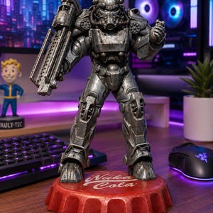May include: A detailed figurine of a soldier in weathered metallic power armour, holding a large weapon on the right arm. The figure stands on a circular red base shaped like a bottle cap with the white text Nuka Cola printed on the top surface.