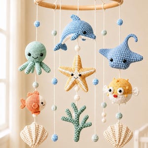 Könnte beinhalten: Ein hängendes Mobile mit gehäkelten Meerestieren, darunter ein blauer Delfin, ein grüner Oktopus, ein gelber Seestern, ein blauer Rochen, ein orangefarbener Fisch, ein gelber Kugelfisch und zwei beige Muscheln, befestigt an einem runden Holzrahmen.