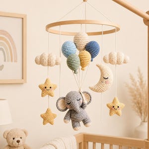 May include: A crochet baby mobile featuring a gray elephant hanging from a cluster of five colorful balloons. The mobile includes two white clouds, two yellow stars with smiling faces, and a crescent moon with a sleeping face, all suspended from a wooden ring.