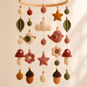 May include: A handmade crochet baby mobile featuring a wooden ring and hanging crochet elements. The mobile includes various shapes like stars, clouds, leaves, birds, flowers, mushrooms, and acorns in shades of cream, pink, orange, and green.