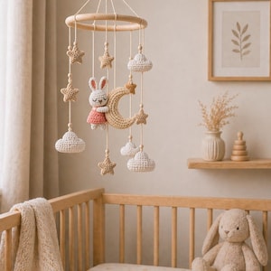 May include: A handmade baby mobile featuring a wooden ring with hanging crochet stars, clouds, and a bunny wearing a pink dress, and a crescent moon. The mobile hangs above a wooden crib with a beige blanket and a stuffed bunny.