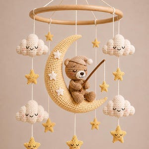 Pode incluir: Um móbile de crochê para bebê com um urso marrom claro usando uma touca de dormir, sentado em uma grande lua crescente amarela. O móbile inclui várias nuvens brancas e estrelas amarelas suspensas em uma estrutura circular de madeira.
