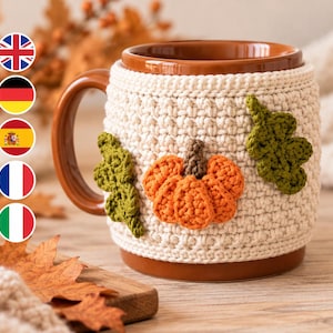 Puede incluir: Una taza de cerámica marrón con una funda de ganchillo color crema. La funda está decorada con una calabaza de ganchillo naranja en el centro con un tallo marrón, flanqueada por dos hojas de roble verdes de ganchillo a cada lado.