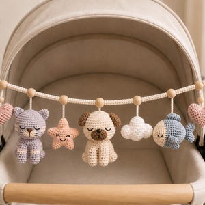 May include: A crochet stroller toy arch featuring a cream-colored cord with wooden beads and hanging figures including two pink hearts, a lavender cat, a pink star, a beige pug, a white cloud, and a blue fish, all suspended from wooden rings.