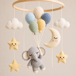Könnte beinhalten: Handgefertigtes Baby-Mobile aus Häkelarbeit mit einem grauen Elefanten, der bunte Luftballons hält. Das Mobile umfasst einen Halbmond, Sterne und Wolken, alles in sanften Pastellfarben, aufgehängt an einem Holzreifen.