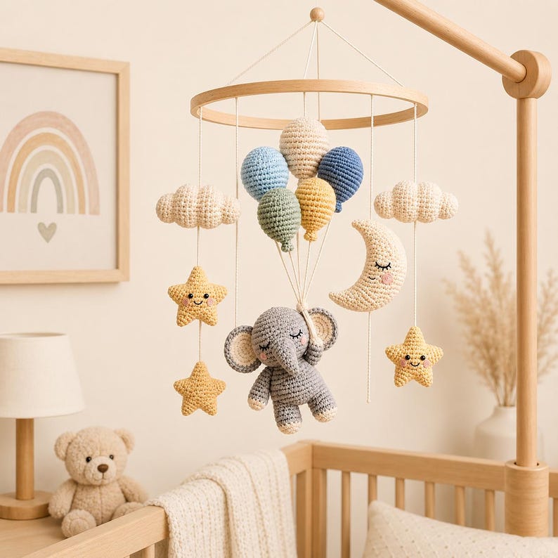 May include: A crochet baby mobile featuring a gray elephant hanging from a cluster of five colorful balloons. The mobile includes two white clouds, two yellow stars with smiling faces, and a crescent moon with a sleeping face, all suspended from a wooden ring.