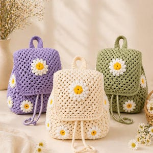May include: Three crochet backpacks in lavender, cream, and green, each adorned with daisy appliqués. The bags have a square shape, drawstring closure, and a top handle. The daisy details feature yellow centers and white petals.