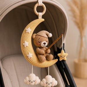 May include: A crochet hanging mobile featuring a yellow crescent moon with small white stars, a brown bear wearing a nightcap, and two white clouds. The bear holds a fishing rod with a yellow star attached. It hangs from a wooden ring.